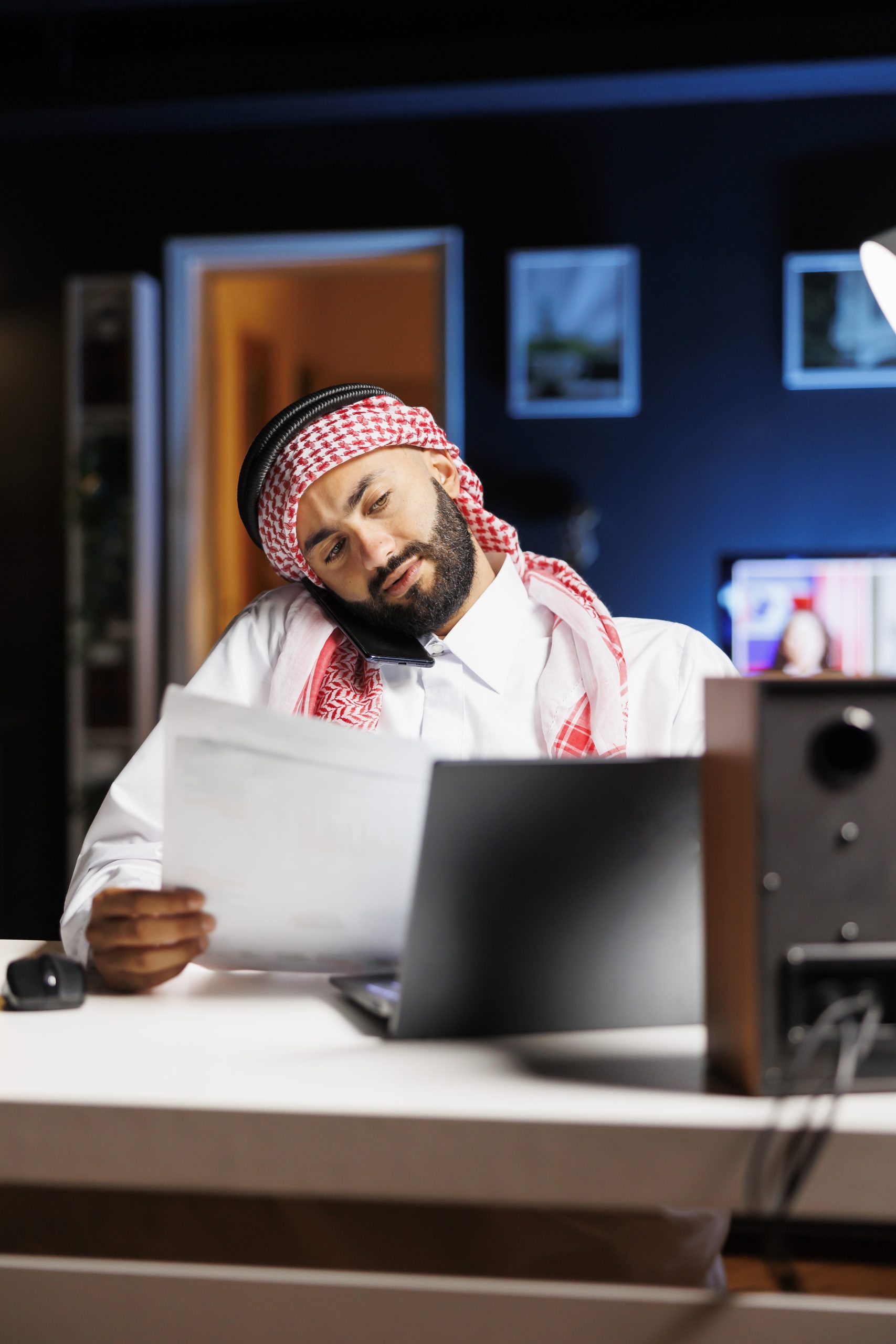 Detailed shot of Arab man using a laptop and mobile device for communication and research, showcasing proficiency in technology. Muslim guy multitasking, talking on his cellphone and comparing notes.